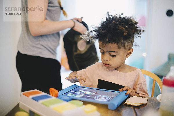 Junge mit digitalem Tablett am Tisch  während die Mutter im Hintergrund steht.
