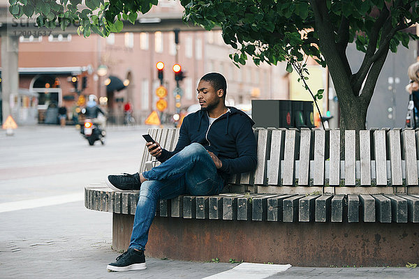 Junger Mann mit Smartphone im Sitzen mit gekreuzten Beinen auf der Bank in der Stadt
