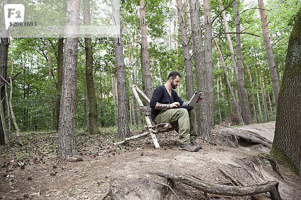 Mann sitzend auf selbstgemachtem Holzstuhl im Wald mit Laptop