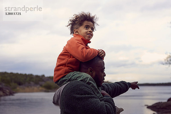 Vater zeigt auf das Meer  während er seinen Sohn auf den Schultern gegen den Himmel trägt.