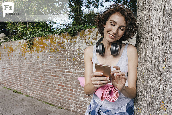 Lächelnde Frau mit Matte  Kopfhörer und Handy auf dem Bürgersteig