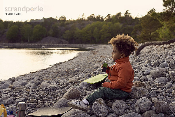 Seitenansicht des Jungen beim Sitzen auf Felsen am Strand bei Sonnenuntergang