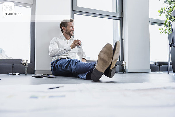 Entspannter Geschäftsmann sitzt auf dem Boden seines Büros bei einer Tasse Kaffee.
