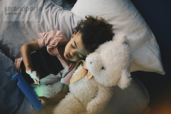 Hochwinkelansicht des Jungen  der mit Teddybär liegt  während er zu Hause mit einem digitalen Tablett auf dem Bett liegt.