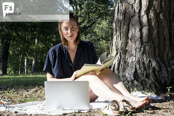 Junge Frau sitzend unter einem Baum mit einem Buch und einem Laptop