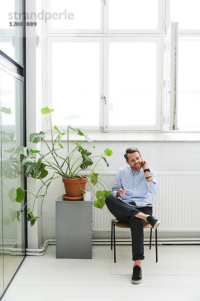 Volle Länge des Geschäftsmannes  der auf dem Handy spricht  während er bei einer Topfpflanze im Kreativbüro sitzt.