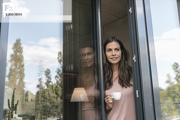 Lächelnde Frau hält Tasse mit Blick aus der Balkontür