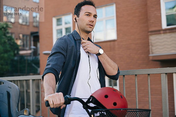 Mittlerer Erwachsener Mann  der In-Ear-Kopfhörer anpasst  während er mit dem Fahrrad gegen das Bauen in der Stadt geht.