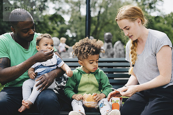 Eltern mit Kindern beim Essen auf der Bank im Park