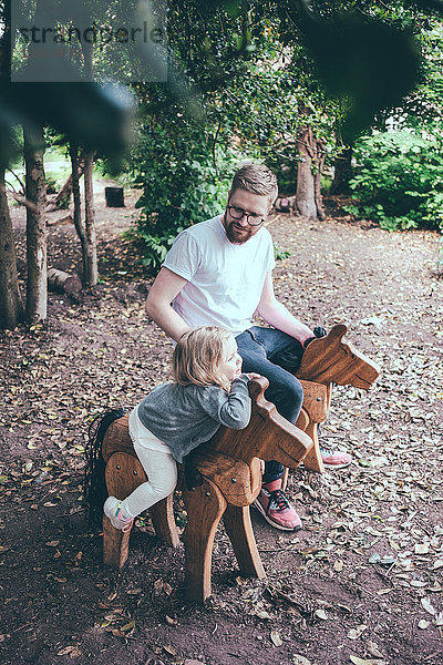 Hochwinkelansicht von Vater und Tochter auf Holzpferden sitzend im Park