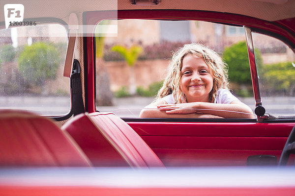 Porträt eines lächelnden blonden Mädchens  das sich an die Autoscheibe lehnt und nach innen schaut.