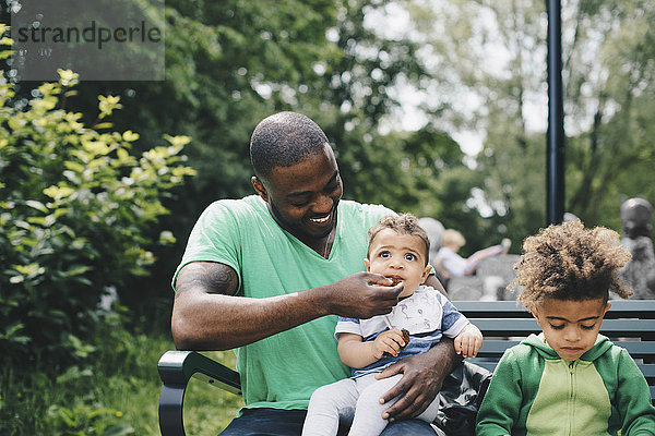 Lächelnder Vater füttert den Jungen  während er mit seinem Sohn auf der Bank im Park sitzt.