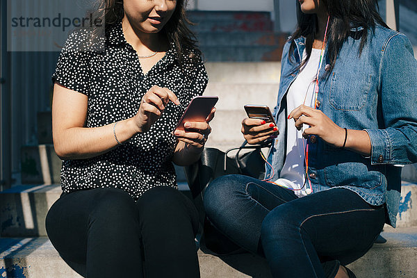 Mittelteil der weiblichen Freunde  die Smartphones benutzen  während sie auf Stufen sitzen.