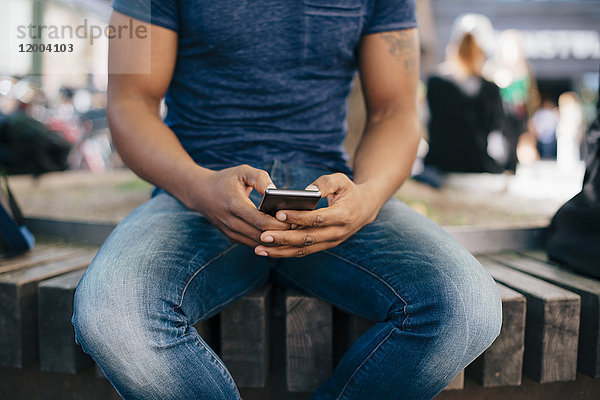 Mittelteil eines jungen Mannes  der ein Smartphone benutzt  während er auf einer Bank in der Stadt sitzt.