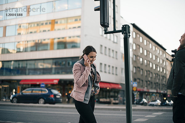 Lächelnde junge Frau  die auf dem Handy spricht und gegen das Gebäude in der Stadt geht.
