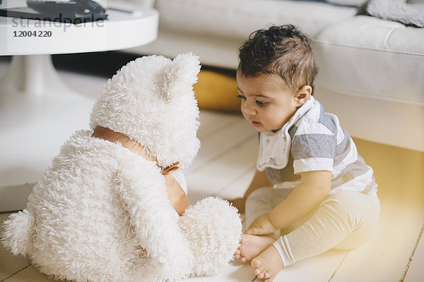 Kleinkind schaut auf den Teddybär  während es zu Hause auf dem Boden sitzt.