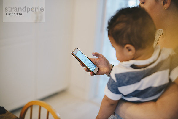 Mutter  die das Handy benutzt  während sie ihren Sohn zu Hause trägt.