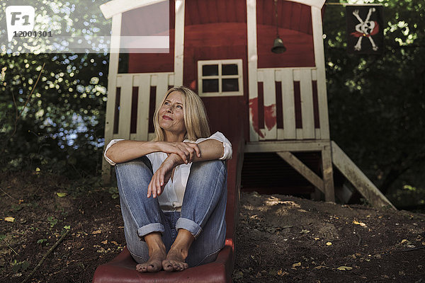 Reife Frau sitzt auf einer Rutsche vor einem Gartenhaus im Wald.