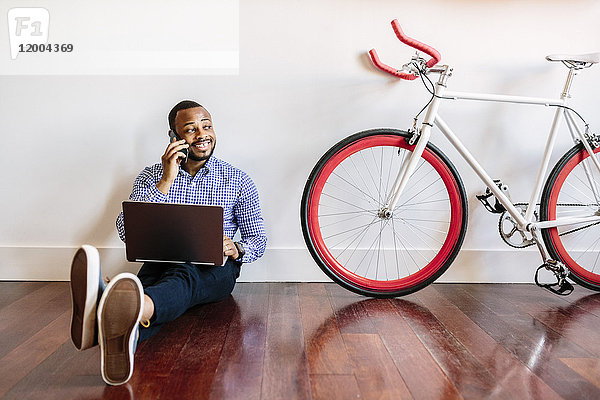 Lächelnder Mann am Handy auf Holzboden sitzend mit Fahrrad neben ihm
