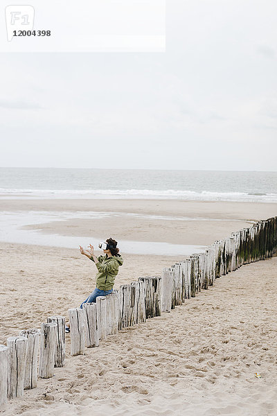 Frau auf Holzpfahl am Strand sitzend mit VR-Brille