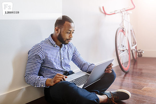 Mann mit Laptop auf Holzboden sitzend mit Fahrrad im Hintergrund