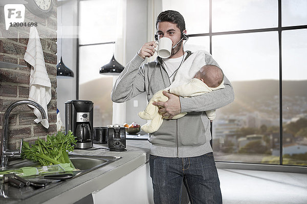 Vater mit Kopfhörer trinkt Kaffee in der Küche hält Baby