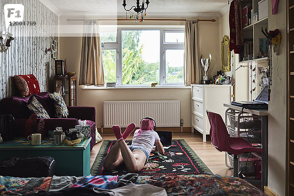Junge Frau mit rosa Haaren  die auf einem Teppich liegt  trägt Kopfhörer und benutzt den Laptop zu Hause.