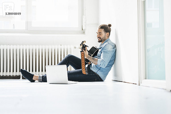 Mann sitzt mit Laptop auf dem Boden und spielt Gitarre
