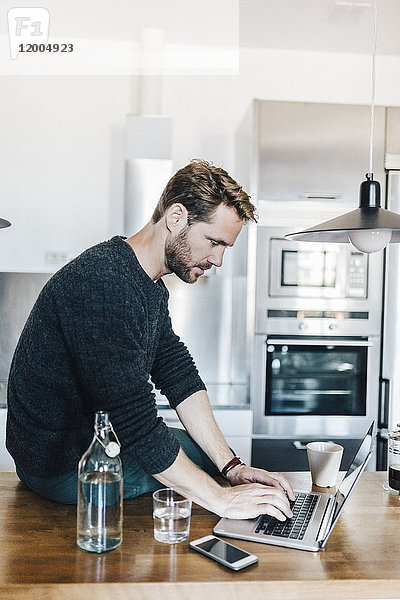 Mann sitzt auf der Arbeitsplatte in der Küche mit Laptop