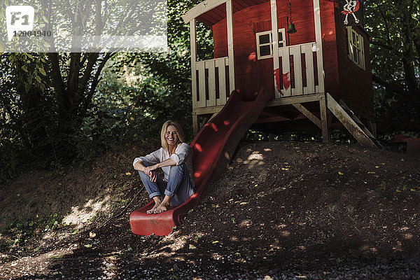 Reife Frau sitzt auf einer Rutsche vor einem Gartenhaus im Wald.