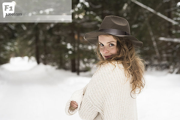 Porträt einer lächelnden jungen Frau mit Hut und Strickpullover im Winterwald