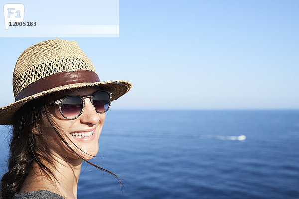 Porträt einer lächelnden Frau mit Strohhut und Sonnenbrille vor dem Meer