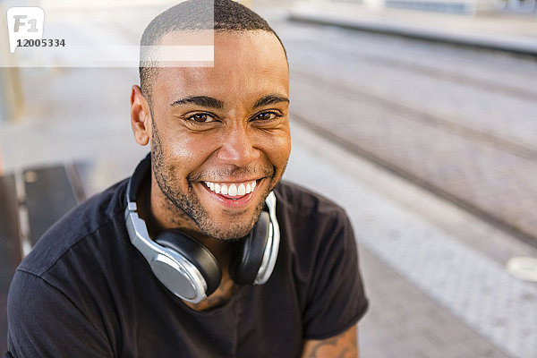 Porträt eines lachenden jungen Mannes mit Kopfhörern an der Straßenbahnhaltestelle