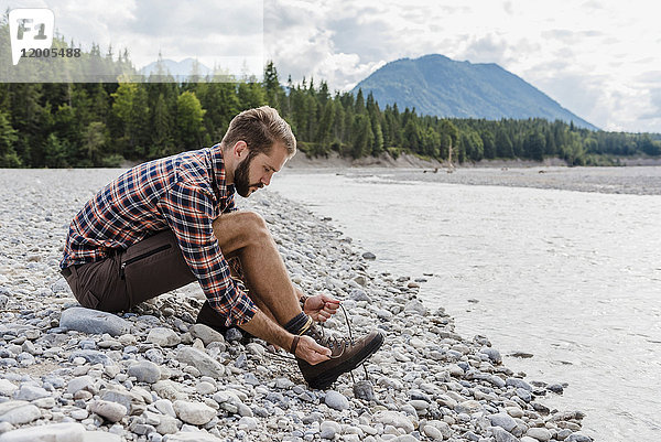 Deutschland  Bayern  Wanderer sitzt am Flussufer und bindet sich die Schuhe.
