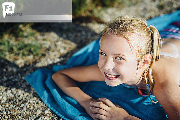 Porträt eines lächelnden  blonden Mädchens auf einem Handtuch am Strand.