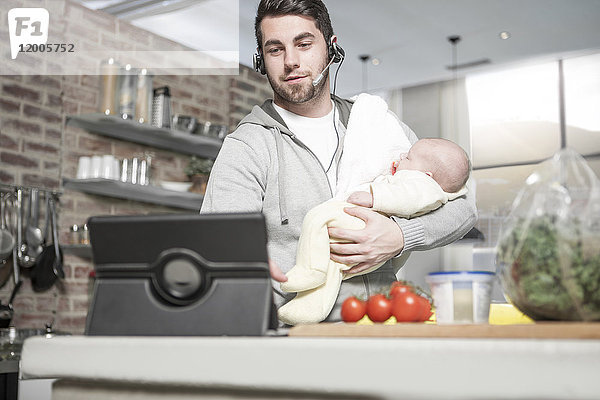 Vater mit Kopfhörer und Tablette in der Küche hält Baby