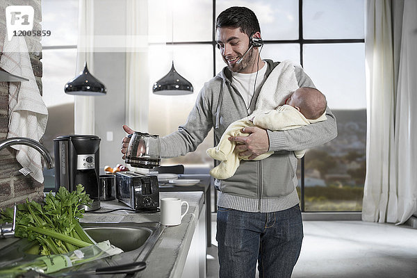 Vater mit Kopfhörer macht Kaffee in der Küche und hält Baby