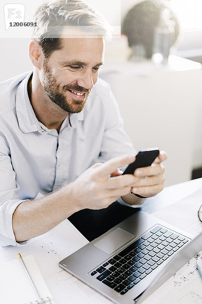 Porträt eines lächelnden Geschäftsmannes mit Smartphone am Schreibtisch in seinem Büro