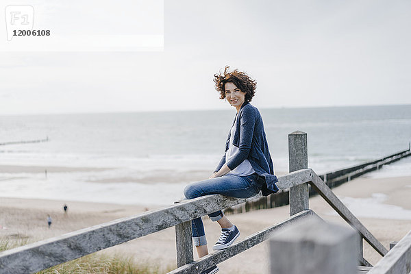 Porträt einer lächelnden Frau  die auf einem Geländer am Strand sitzt.