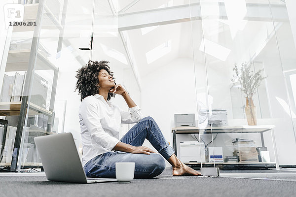 Junge Frau mit geschlossenen Augen sitzend auf dem Boden im Büro mit Laptop