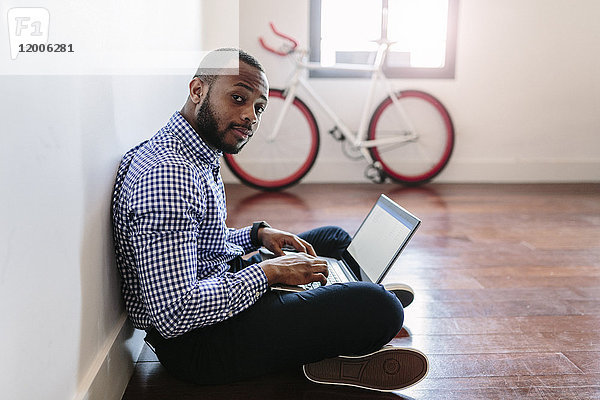 Mann mit Laptop auf Holzboden sitzend mit Fahrrad im Hintergrund
