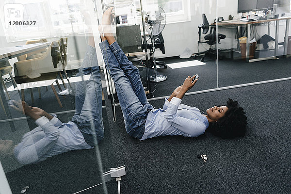 Junge Frau  die im Büro auf dem Boden liegt und ihr Handy überprüft.