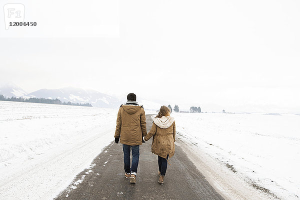 Rückansicht eines verliebten jungen Paares  das Hand in Hand auf einer Landstraße in einer verschneiten Landschaft wandert.