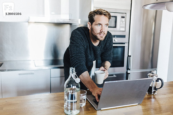 Portrait des Hauptdarstellers mit Kaffeetasse und Laptop in der Küche stehend