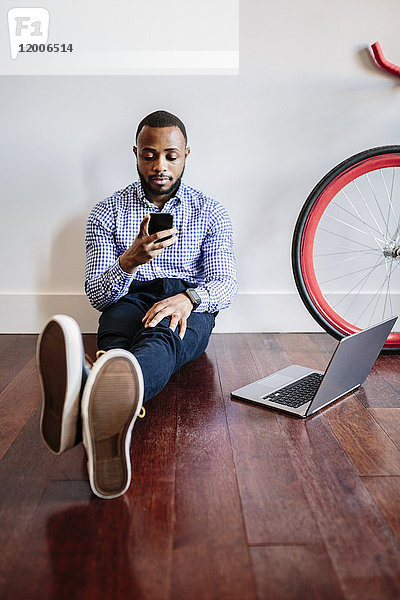 Mann auf Holzboden sitzend mit Laptop und Handy und Fahrrad neben ihm