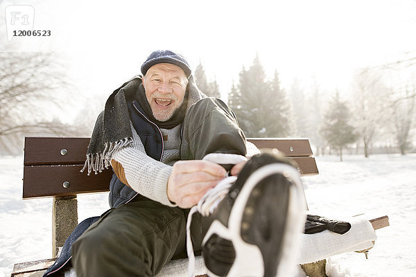 Porträt eines glücklichen älteren Mannes  der auf einer Bank in der Winterlandschaft sitzt und Schlittschuhe anzieht.