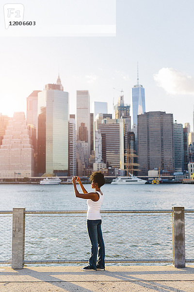USA  New York City  Brooklyn  Frau steht am Wasser und macht Handyfotos