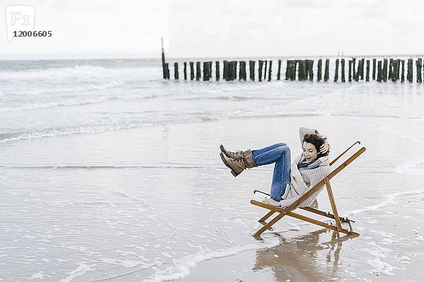 Frau sitzt auf einem Liegestuhl am Strand und hebt ihre Beine.