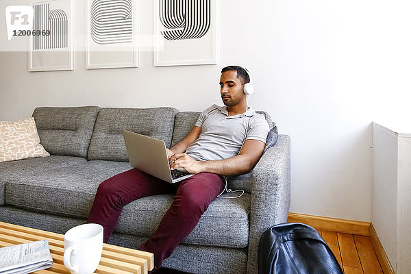 Gemischtrassiger Mann sitzt auf dem Sofa und hört mit Kopfhörern auf seinen Laptop