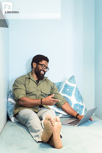 Lächelnder schwarzer Mann sitzt auf einem Bett und zeigt auf einen Laptop